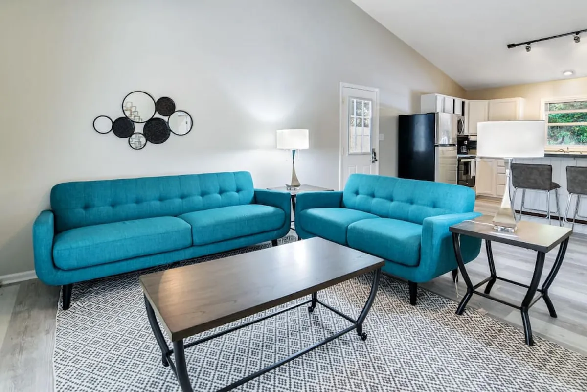 The hero image showcases a stylish, open-concept living area with inviting turquoise sofas, a sleek black coffee table atop a geometric rug, and modern decorative wall mirrors. The kitchen is visible in the background, enhancing the sense of space and sociability. The atmosphere is both contemporary and cozy, perfect for relaxing, socializing, or enjoying quiet moments.