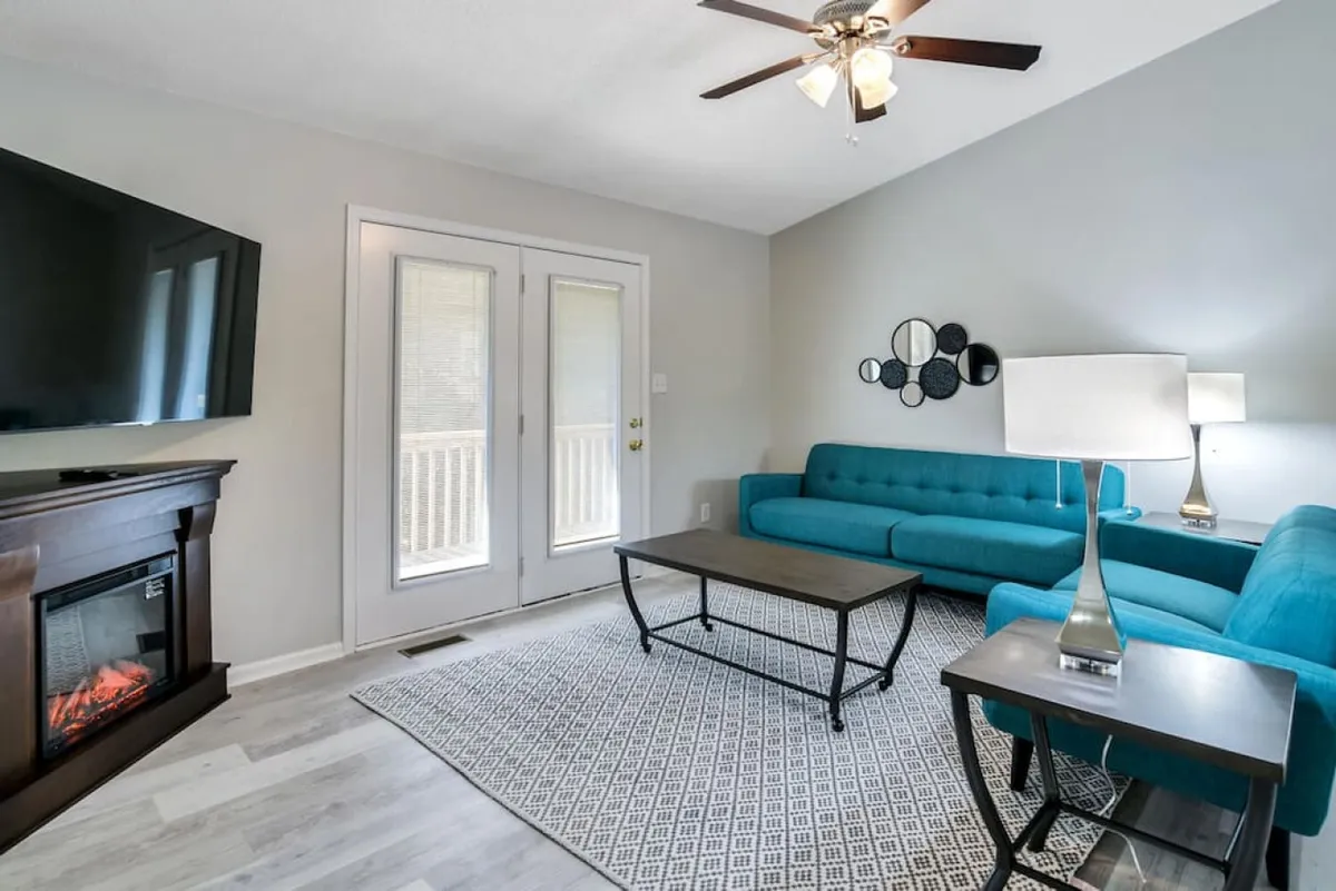 A bright and open living room featuring a stylish turquoise sofa, a modern coffee table, a cozy fireplace, and a TV. Double doors let in streams of natural light, highlighting the contemporary furnishings and creating an inviting space for relaxation or socializing.