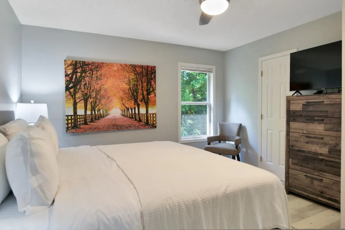 A bright, peaceful bedroom anchored by a bed with crisp white linens and a large window letting in abundant natural light. A striking piece of artwork depicting a sun-dappled, tree-lined path hangs above the bed, while a cozy chair in the corner invites relaxation or reading. Soft shadows and greenery glimpsed outside reinforce the tranquil vibe.