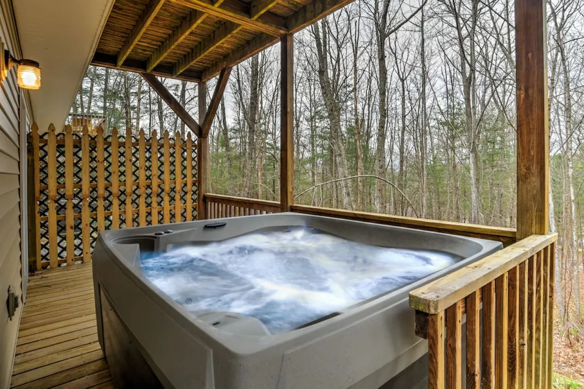 This image features a serene outdoor deck nestled amidst lush trees, showcasing a hot tub as the focal point. The wooden deck provides ample space for relaxing or socializing, while the surrounding greenery conveys a sense of privacy and natural beauty—ideal for enjoying morning coffee or evening drinks outdoors.