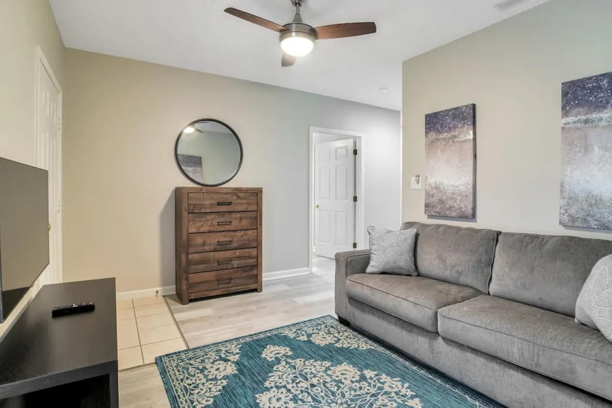 A spacious and welcoming living area featuring a cozy sofa, a patterned area rug, a mirror above a dresser, and decorative artwork. The space exudes warmth and is ideal for entertaining, conversation, or relaxing with a pet.