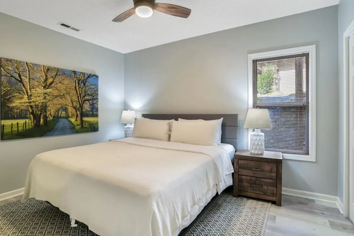 A cozy bedroom scene with a comfortable bed dressed in white bedding, flanked by nightstands and lamps. Tree-themed wall art adds a touch of nature-inspired décor, and sunlight gently filters through a nearby window. The room offers both warmth and tranquility, perfect for unwinding after an adventurous day.