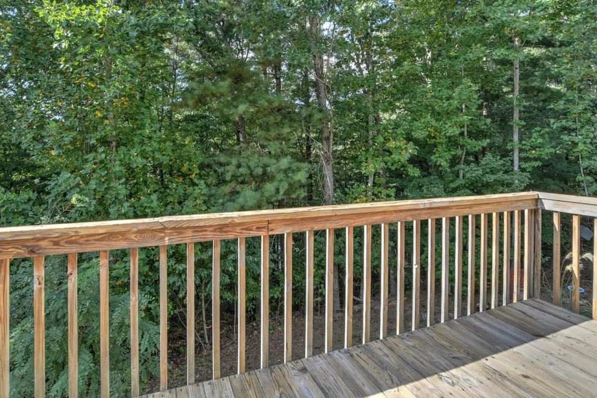 This exterior image shows a peaceful deck with a rustic wooden railing, surrounded by dense greenery. The deck surface is bathed in natural light, offering a serene space that overlooks nature, ideal for relaxing or enjoying the outdoors with pets.